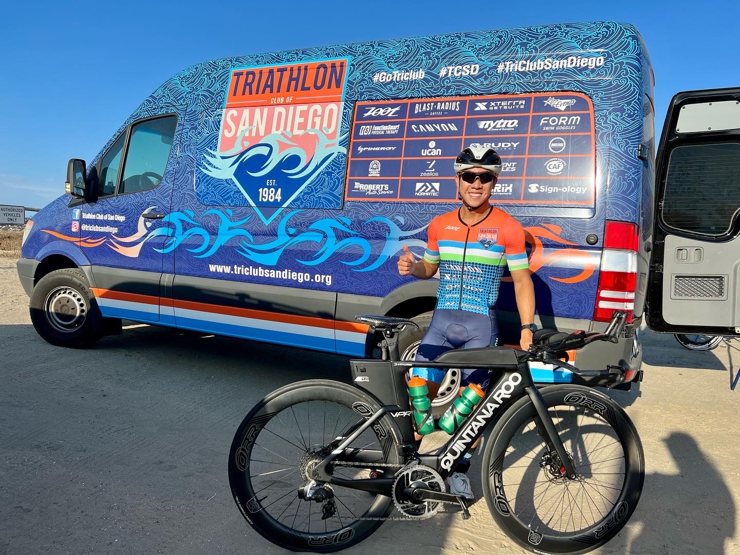 Anthony Chan in his Triathlon Club of San Diego kit with his Quintana Roo bike beside the TCSD team van