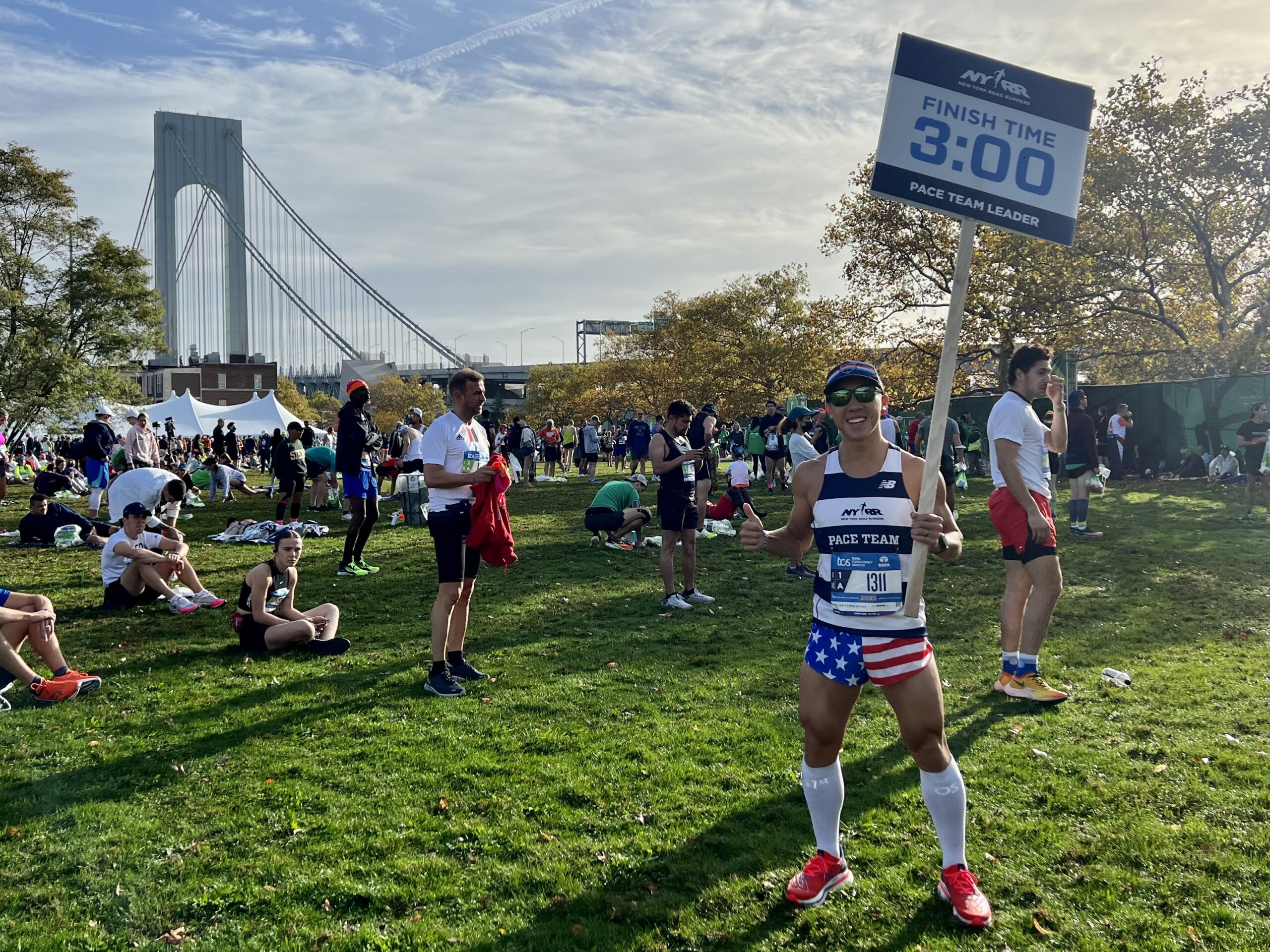 Anthony Chan leading the 3:00 pace team at the New York City Marathon, holding the pace sign with the Verrazzano Bridge behind him